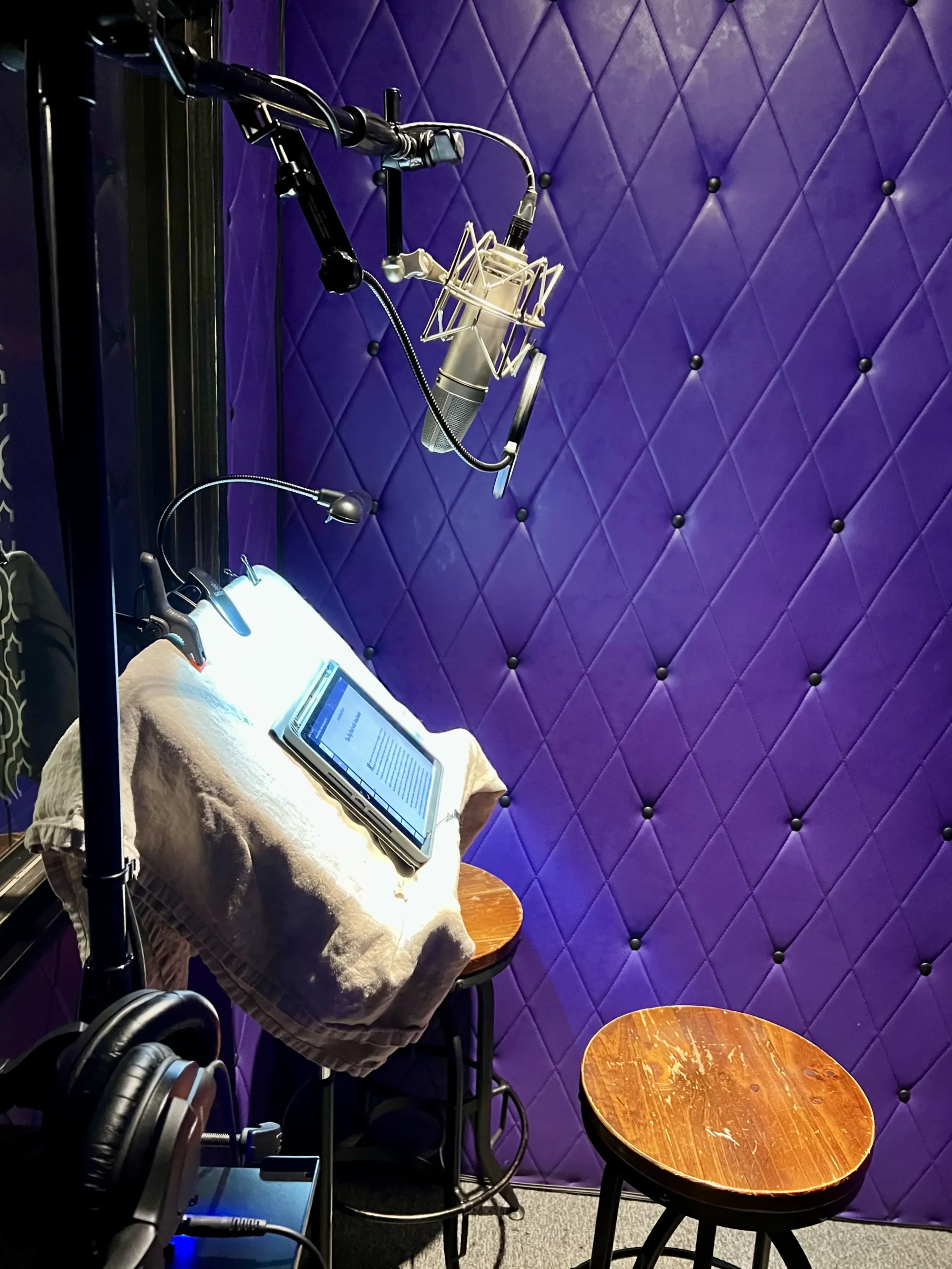 Recording studio setup with a microphone, pop filter, headphones, and a digital tablet on a music stand, against a purple padded wall.