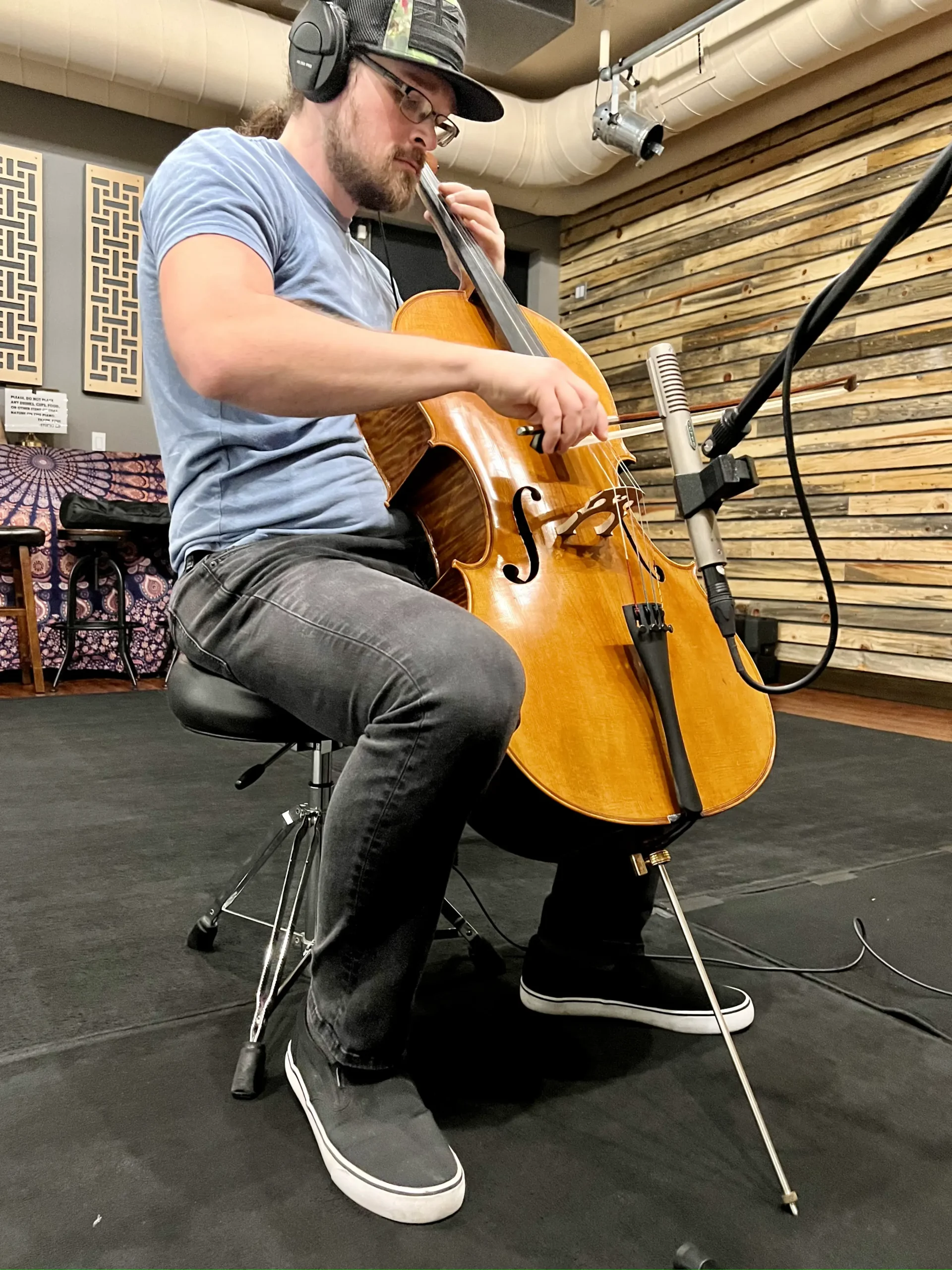 A man in a studio wearing headphones plays a cello, positioned in front of a microphone, with a focused expression.