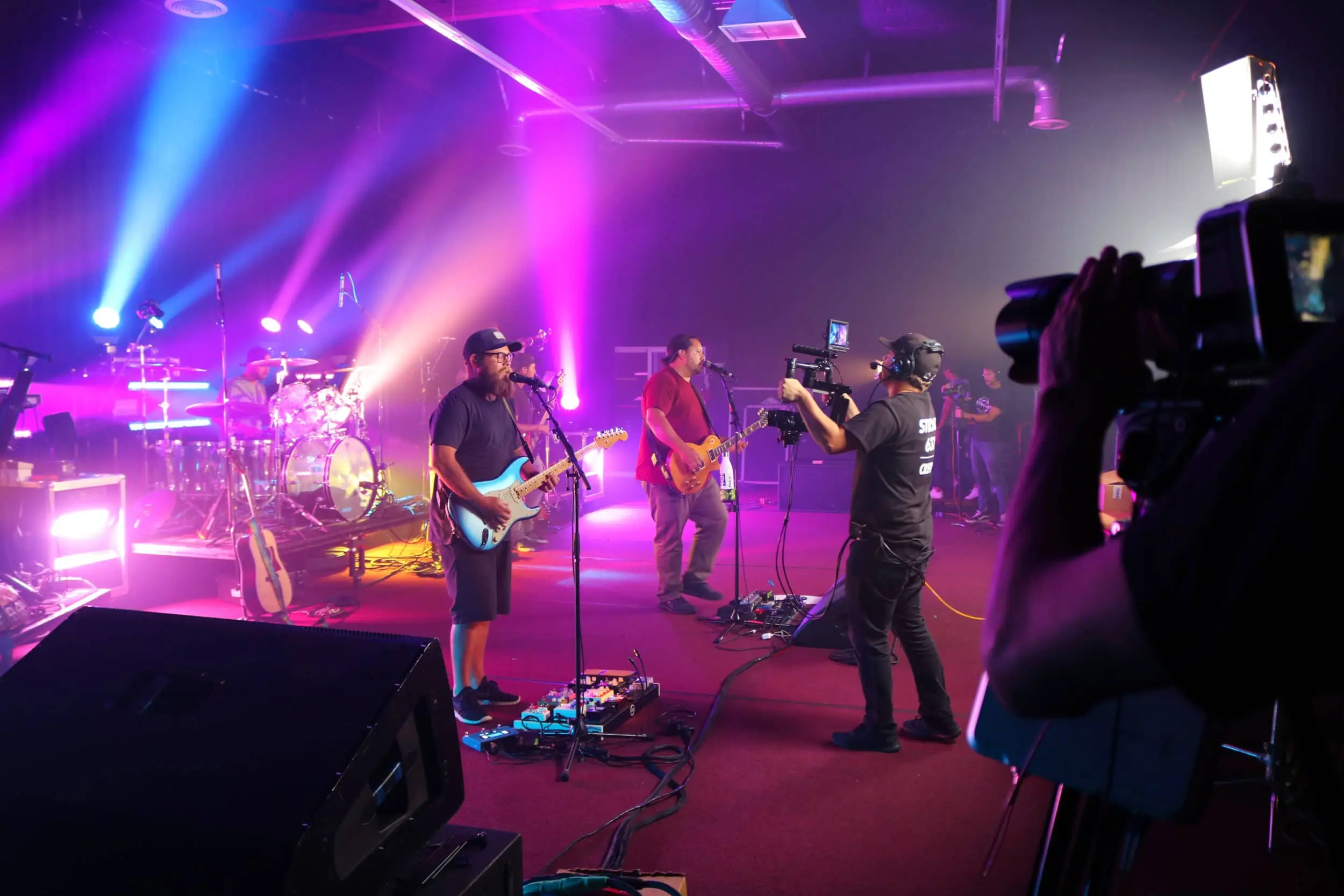 A band performs on stage under pink lighting while a cameraman records the event.