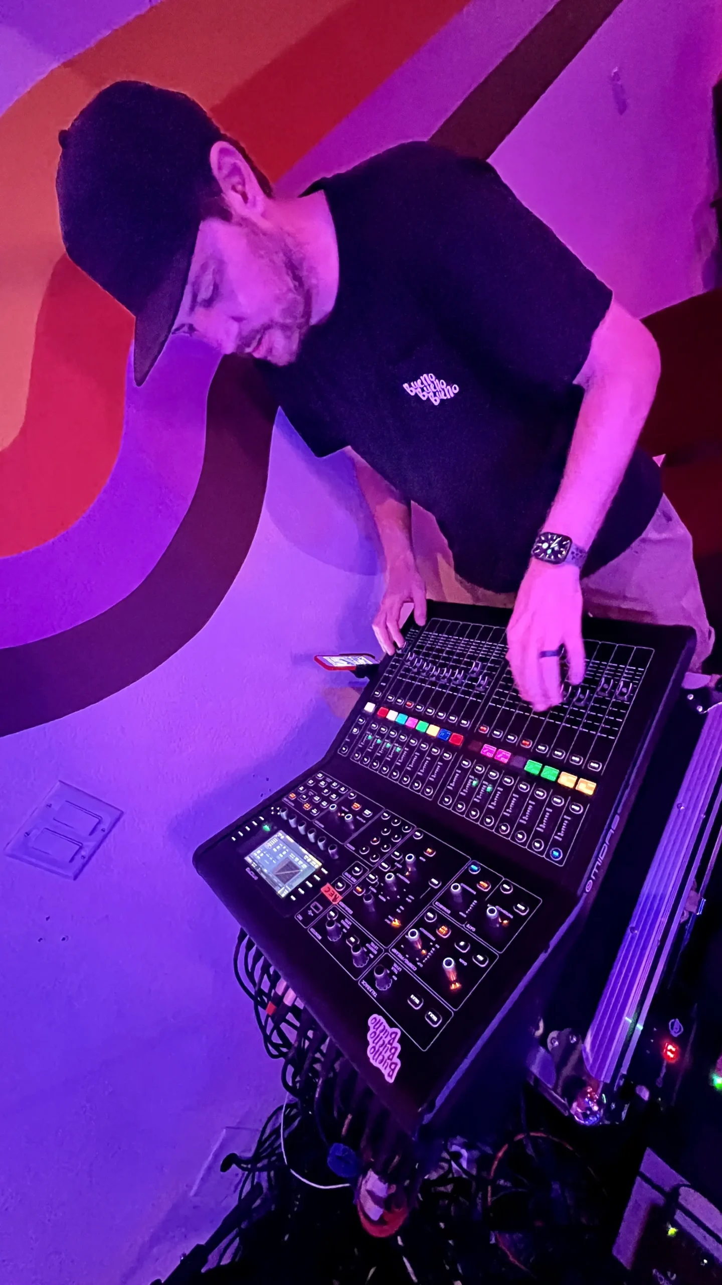 A man adjusting controls on a large electronic music mixer, lit by purple ambient lighting. he wears a cap and a black shirt.