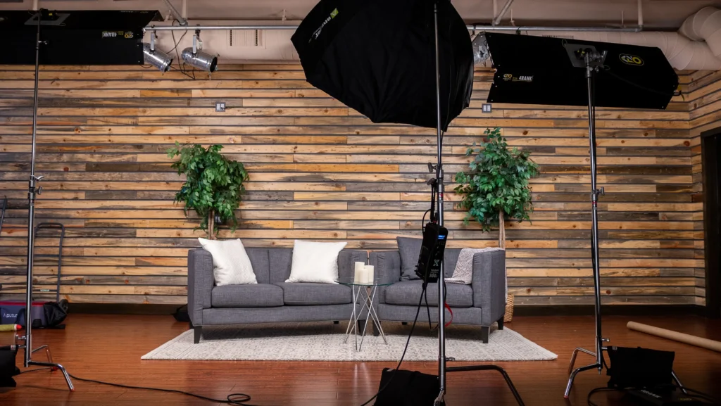 A professional video or photo studio setup with a gray couch, two green plants, lighting equipment, and a camera on a tripod.
